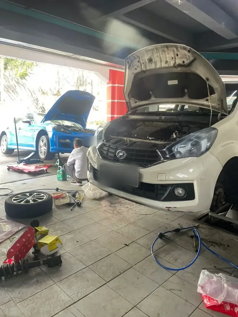 White hatchback with its bonnet raised in the foreground and a blue car also open behind it, a removed wheel and tools on the floor