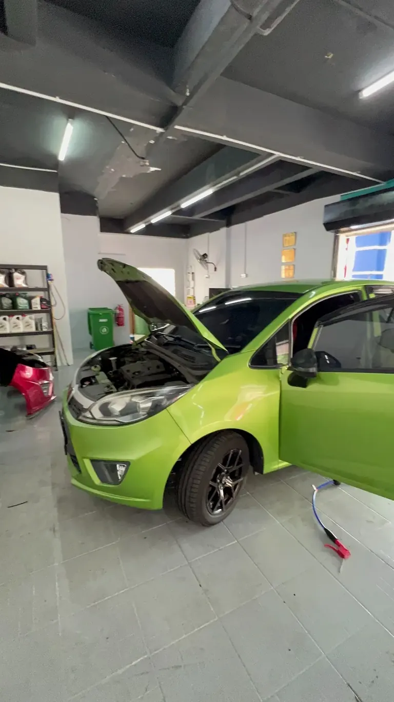 Green hatchback with bonnet and door open inside the workshop, shelves of fluids at the left