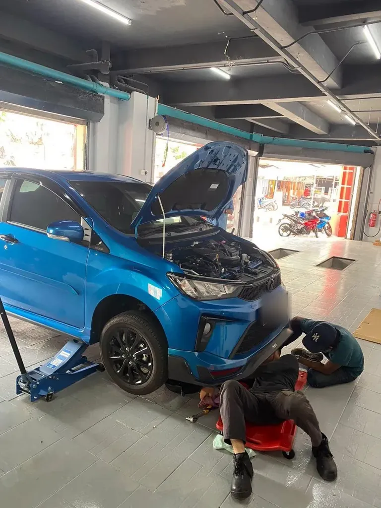Blue hatchback jacked up with its bonnet open, one technician under the front on a creeper and another beside the wheel