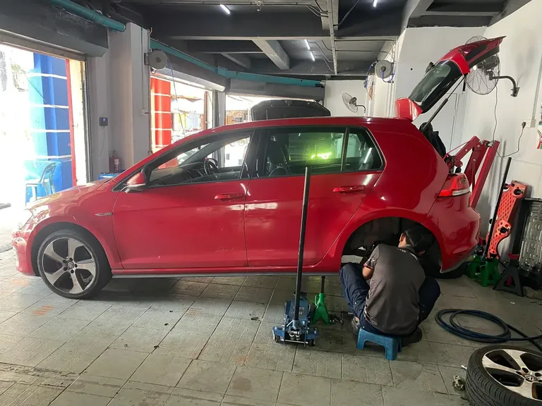 Red hatchback raised on a trolley jack while a technician works at the rear wheel